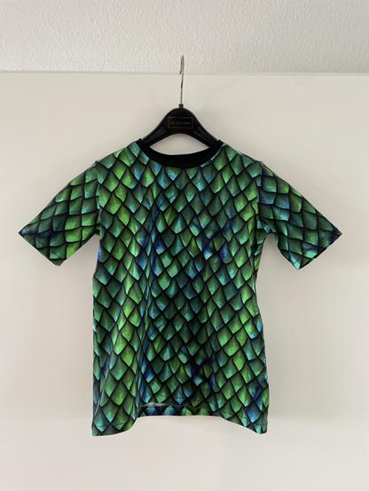 Auf einem Kleiderbügel an einer Zimmertür hängt ein Kinder-T-Shirt aus grün-blauem Drachenschuppen-Jersey mit schwarzem Halsbündchen.