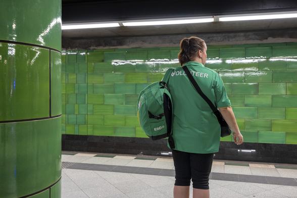 A lady dressed in a green short sleeves shirt, black shorts and a green backpack is waiting for her train in a metro station in Berlin decorated with shiny green tiles both on the wall and on a central pillar.