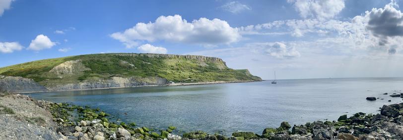 A panoramic view of a coastal landscape featuring a grassy cliff, calm water, and a sailboat in the distance. The sky is partly cloudy, with some visible rock formations and patches of green moss along the shoreline.
