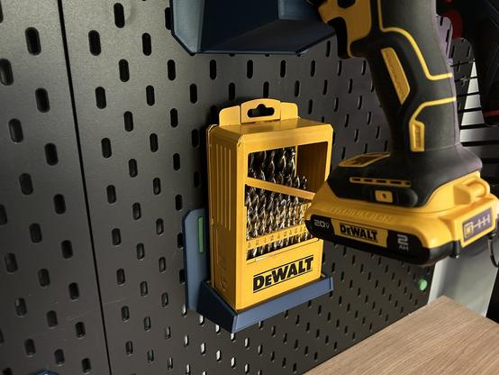 A 3D printed shelf hangs on an IKEA SKÅDIS pegboard.  A DeWalt drill index (metal box of bits) sits in the cavity in the shelf.  The whole thing is behind the handle of a DeWalt cordless drill, which is hanging on a bracket just above.