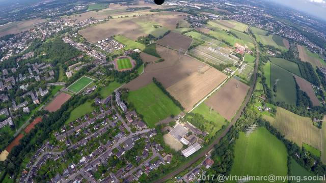 An aerial photo mostly of the no mans land between Essen and Bochum with big farm yards and a big Electrical substation. There are living quaters in the green with trees all around at the bottom. In the distance the city Bochum can be seen at the top.