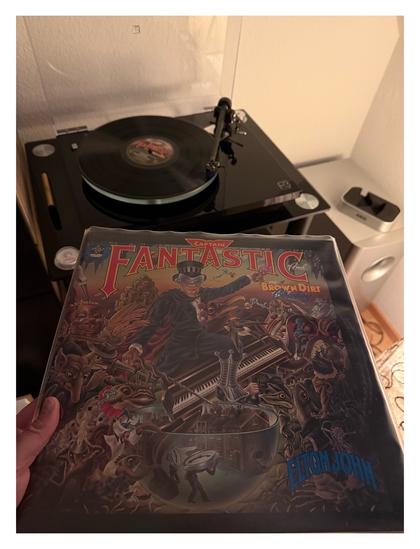 Auf einem schwarzen Plattenspieler läuft eine schwarze Schallplatte. Davor halte ich das Cover ins Bild. Eine gezeichnete Renaissance artige Collage der unterschiedlichsten Dinge, mittig reitet Elton John, bombastisch gekleidet, auf einem Klavier.