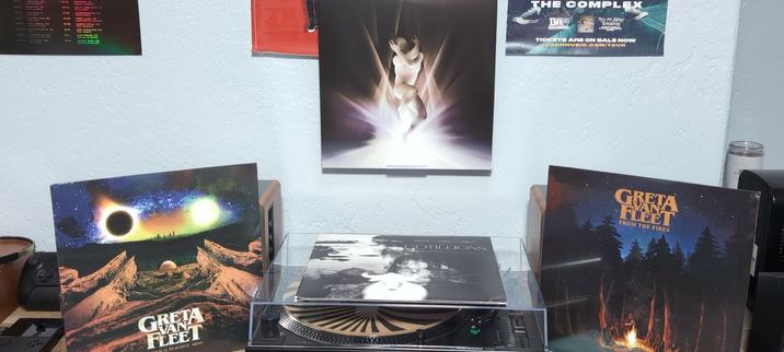 The image shows a collection of vinyl records and posters on a wall. In the center, there is a turntable with an album laying on it.. To the left, there is an album cover for "Greta Van Fleet" featuring a cosmic scene with a solar eclipse and a mountainous landscape. To the right, another album cover for "Greta Van Fleet" titled "From the Fires" depicts a forest scene with a campfire. Above the turntable, there is an album by The Smashing Pumpkins. On the left side of the image, there is a partial view of a poster, and on the right, a poster advertising "The Complex" with information about ticket sales. The wall is white, and the overall setting appears to be a personal music collection area.