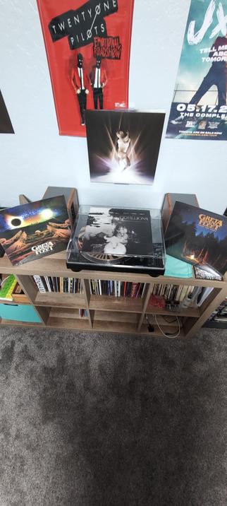 The image shows a wooden bookshelf with a turntable on top, surrounded by various vinyl records and posters. The shelf is filled with CDs and records, with a prominent turntable in the center. On the left, there is a vinyl record cover titled "Greta Van Fleet" with an image of a solar eclipse. In the center, a record titled "Cotillions" by William Patrick Corgan is visible. On the right, another "Greta Van Fleet" record is seen, featuring a campfire scene. Above the shelf, two posters are visible: one for "Twenty One Pilots" with the album "Emotional Roadshow," and another for "jxdn" with the album "Tell Me About Tomorrow." The wall behind the shelf is light-colored, and the floor is covered with a gray carpet.
