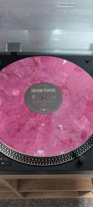 The image shows a pink marbled vinyl record on a turntable. The record is labeled "SMASHING PUMPKINS" with the text "THE COLOUR OF LOVE" and "CONFESSIONS OF A DOPAMINE ADDICT" on the label. The turntable is black with a silver dot-patterned edge, and the platter has a speed indicator showing "45." The record is placed on the turntable's platter, and the turntable is on a wooden shelf with a textured white wall in the background.