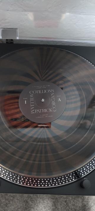 The image shows a vinyl record on a turntable. The record is a transparent black color with a radial pattern of concentric circles. The label in the center reads "COTILLIONS WILLIAM PATRICK CORGAN" and lists the track titles: "1. TO SCATTER ONE'S OWN," "2. HARD TIMES," "3. JUBILEE," and "4. FRAGILE, THE SPARK." The record is labeled as "DISC 1 SIDE A." The turntable has a black base with a perforated edge, and a small knob is visible on the right side. The background is a light-colored wall with a textured surface, and a reflection of a person's hand is visible on the turntable's lid.