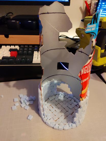 The image shows a creative project made from a Pringles can. The can has been cut and shaped to resemble a castle-like structure. The top part of the can is cut out, revealing a hollow interior, and the sides have been modified to create openings and a textured surface. The bottom of the can is covered with small, light blue foam pieces, arranged to mimic a stone foundation. The foam pieces are scattered around the base of the structure, indicating ongoing work.