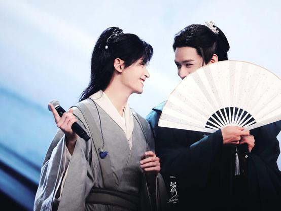 Zhang Zhehan and Gong Jun smiling at each other onstage behind GJ’s fan