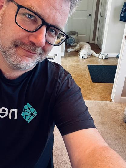 a man with glasses and a beard in the foreground with a dog laying on tile floor behind him.
