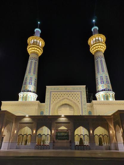 The entrance of the Imam Reza shrine. One the left and right there are two rall round towers, mostly white marble but it has a few colourful parts. On the top of those towers there are some golden parts which are a bit wider than the tower itself.