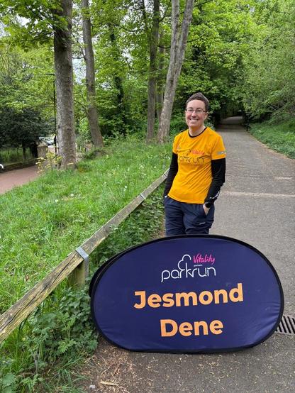 Photo of Emily standing behind a sign that reads “Jesmond Dene parkrun”.