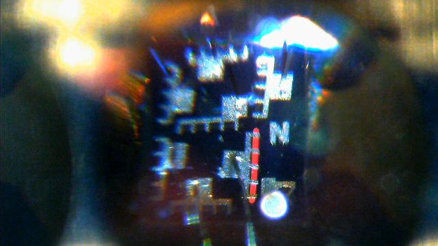 Microscope view of a silicon die, quite distorted due to the "bubble" lens on top of it. 7 fork-like pads made of a silver-colored metal, make up the elements of the 7-segment display. The fork side of the pad has 5 prongs, with the gaps slightly wider than deep. On the bottom right segment, there's a vertical, glowing red line,  which appears to be located under the prongs, extending a bit past the outer-most prongs.