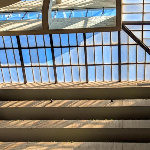 Square color photograph of a skylight and severel floors of balconies. There is sunlight streaming through the skylight adding angles to a very rectangular grid-like image.