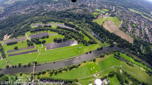 An aerial photo of the ruhr river with some lakes next to it. There are many trees and big grass landscapes next to it. You can see houses in the distance on the top.