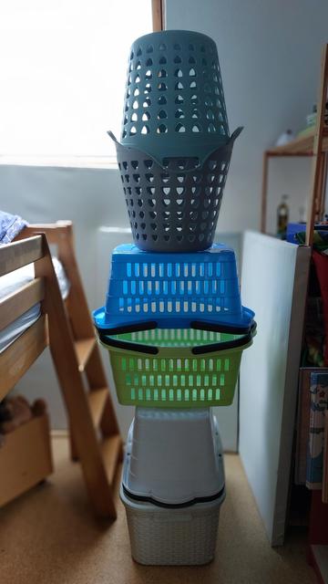 In einem Zimmer stehen sechs leere Wäschekörbe aus Plastik in den Farben weiß, grau, grün und blau. Die Körbe sind so aufeinander gestapelt, dass sie wie eine abstrakte Figur mit Beinen, Rumpf und Kopf aussehen. Links und rechts von dieser Skulptur sind unscharf eine Leiter und ein Regal mit Spielzeug zu sehen, im Hintergrund ein Fenster im Anschnitt.

🇬🇧"Sculpture in the children's room" (laundry baskets misappropriated)
Six empty plastic laundry baskets in white, grey, green and blue. The baskets are stacked on top of each other in such a way that they look like an abstract figure with legs, torso and head. Left and right of this sculpture, a ladder and a shelf with toys can be seen slightly blurred, as well as a window in the background.