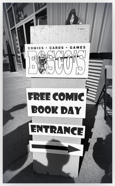 Black and white image of sign on sidewalk  that says : "BOSCOS'S free comic book day entrance" an arrow pointing toward the right under the words