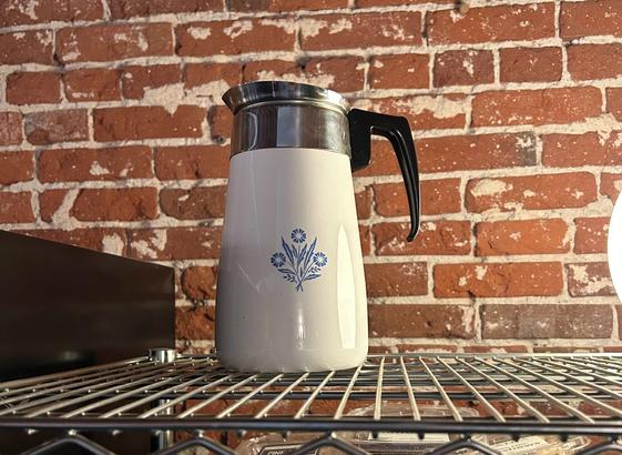 Photo of a vintage Corningware brand blue cornflower design coffee percolator. blue design on white pot, with a black handle and silver top. The pot is displayed on a metal shelf with a bare red brick wall in the background.