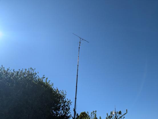 A tall mast with a vertical antenna on top and just below, a 23cm band yagi. The sky is blue and there are trees visible at the bottom of the picture