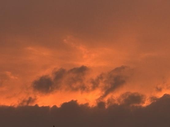 Am unteren Bildrand von links nach rechts eine graue dicke Wolkenwand. Darüber zeigt sich bewölkter Himmel im Hintergrund, der orangerot von der untergehenden Sonne intensiv ausgeleuchtet wird.