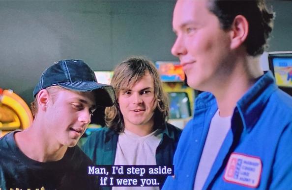 Two young (in early 20s) Jack Black and Giovanni Ribisi in an arcade. Jack has said: "Man, I'd step aside if I were you" to a young man a head taller than them. They are all smiling.