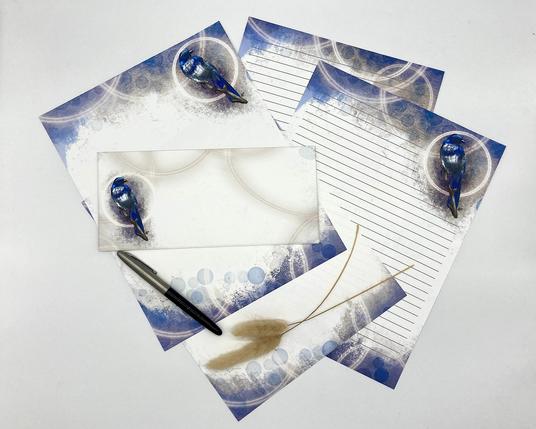 A photograph of multiple sheets of paper stationery with a Steller’s Jay illustration on them. Additional features include blue pastel textures and glowing halos of light on the papers, which are arranged on a white background.