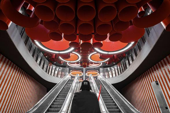 Self-portrait in a Darth Vador costume facing the futuristic background of the Pannenhuis metro station in Belgium, with a giant escalator ascending under a ceiling composed of red and orange giant tubes and lamps hanging.