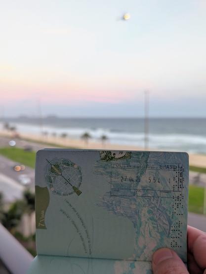 An Irish passport shows an arrival stamp from the Federal Police of Brazil; in the background, the late-afternoon sun lights up Barra di Tijuca's beach while a helicopter flies overhead.