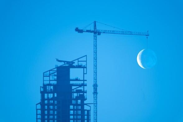 A photograph showing the skeleton of a skyscraper under construction against a blue sky, with a nearby crane turned such that it appears to be raising a crescent moon on its hook.
