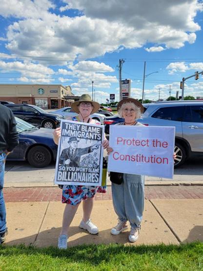 Photo of 2 women standing next to a busy street holding signs. One sign says They Blame Immigrants so you won't blame Billionaires and the 2nd sign says Protect the Constitution