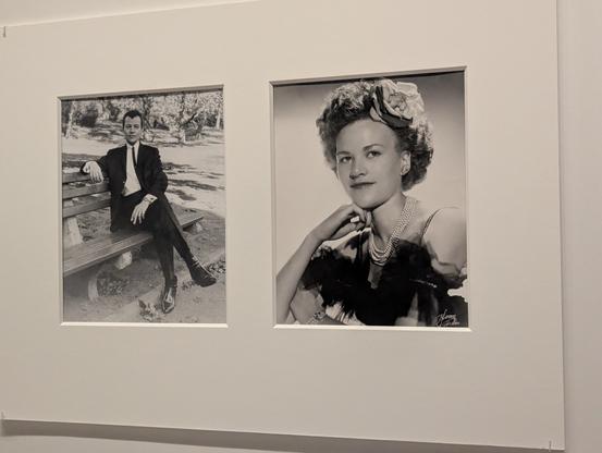 Two black and white photos of the same person, in masc drag (a suit with a thin tie, lounging on a park bench) and in a highly femme outfit (a frilly, off-the-shoulder dress, pearl necklaces, and a flower in the hair).