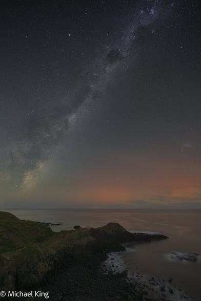The core of the Milky Way rising over the coastline, with an orange glow from Aurora Australis.