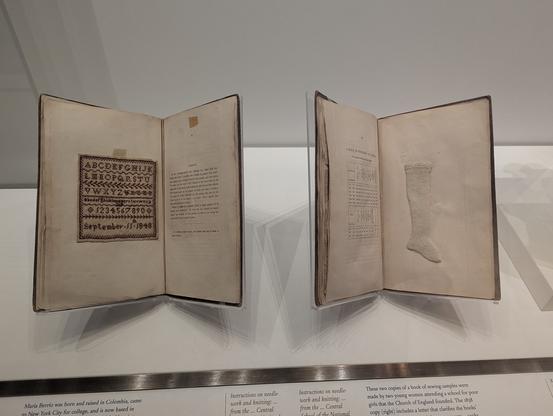 Two books, in stands that hold them open. The one on the right has a tiny knitted stocking. The one on the left has a sampler in red cross stitch, with two alphabets, a row of numbers, and "September 11th 1848".