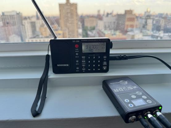 A radio, tuned to 91.7 FM, and a small audio recorder, on a windowsill overlooking an urban skyline.