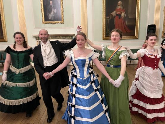 A group of five people dressed in elaborate Victorian-style ballroom attire pose in a formal dance formation in an elegant room with classical portraits and ornate decor. The women wear colorful, full-skirted gowns in green, blue, olive, and burgundy, while the man wears a black tailcoat with a white waistcoat and cravat. They are holding hands, smiling