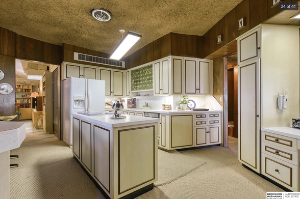 A retro looking white kitchen with brown detailing, and a ceiling that is covered with mustard covered shag carpet.