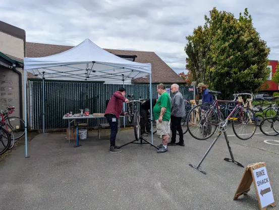 Ouside Shoreline Tool Library. A canopy on the left with a couple of people under it, with a few more people standing to the right and a few used bikes for sale.