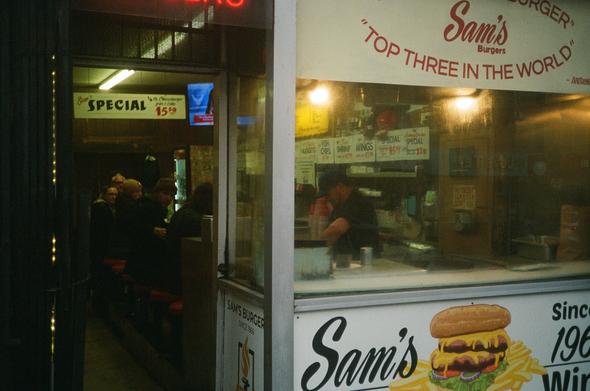 A row of patrons at Sam’s burger quizzically look into the camera or at the TV above the grille.