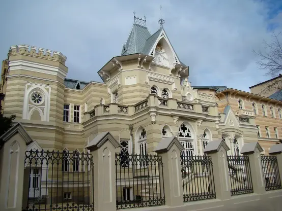 The image depicts a large, ornate building with architectural details typical of a historic or elegant style. It features decorative elements such as turrets, intricate moldings, and large windows. The color palette includes light shades, likely cream or pale yellow, giving it a classic appearance. A wrought iron fence surrounds the property, which adds to its charm. The sky in the background is partly cloudy.

Image Credits: Wikimedia / Giorgi1975 at Georgian Wikipedia / CC BY-SA 3.0