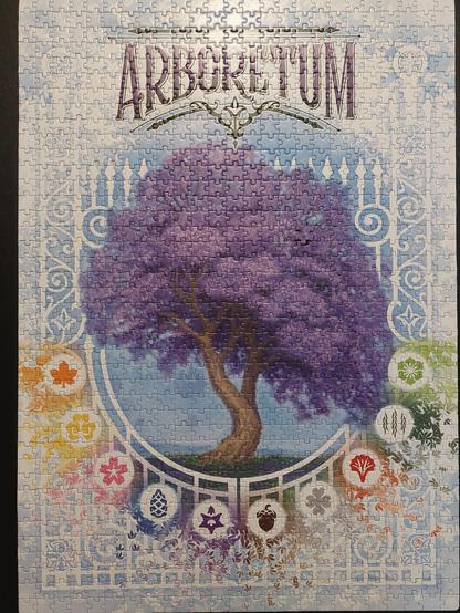 Completed jigsaw puzzle of the game box cover for thr game Arboretum, with a large Jacaranda in the center, game icons in a rainbow underneath, and complicated blue and white patterns elsewhere. One piece does not fit in its spot.