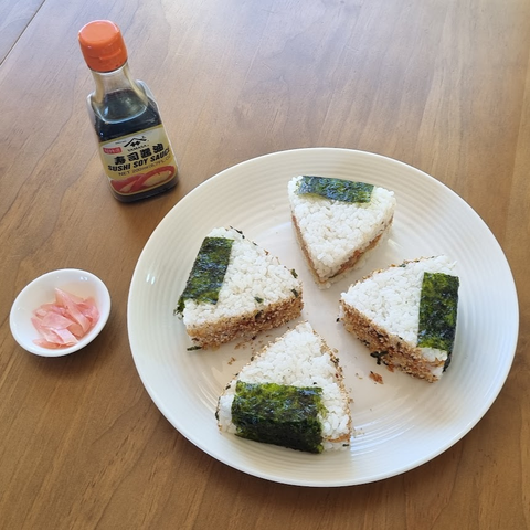 A plate with four onigiri (Japanese rice balls) stuffed with fresh smoked tarakihi with red pepper, fried shallots and avocado, and wrapped in furikake (rice seasoning) and nori (seaweed). Next to the plate is a bottle of soy sauce and some pickled ginger.