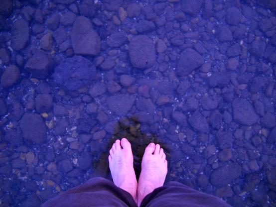 Feet standing on the surface of still clear water with pebbles and rocks visible below the surface