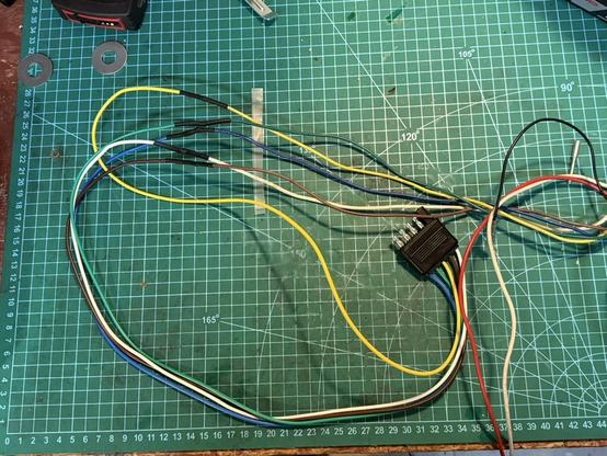 a motorcycle trailer wiring harness on a green project mat. there are long multicoloured wires winding back and forth with shrink-wrapped solder joints in the middle, leading to a 5-pin trailer connector.