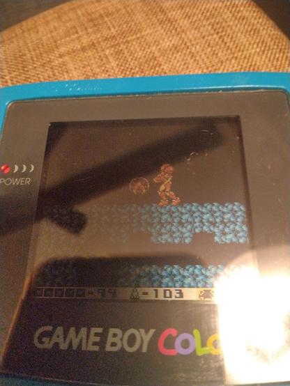 Photo of a Game Boy Color, with Metroid II on the screen, showing Samus with a tiny Metroid.