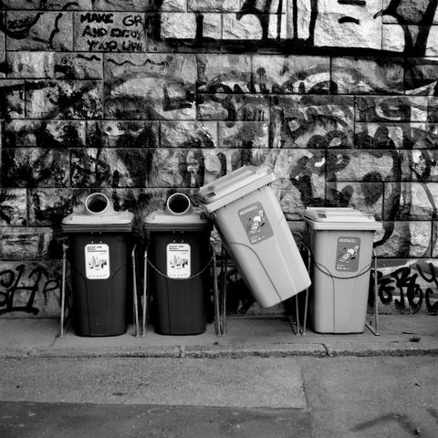 The black and white image shows four trash and recycling bins lined up against a graffiti-covered brick wall, with one of the bins leaning diagonally to the side, creating a sense of imbalance.