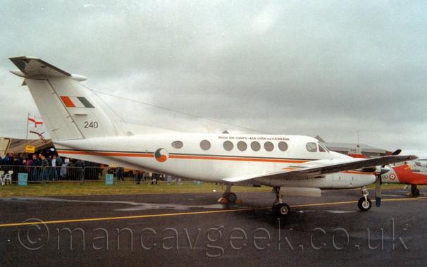 Side view of a white, twin engined Bizprop parked on a black tarmac taxiway facing to the right.
There are small, black "Irish Air Corps" titles on the upper forward fuselage, in Irish at the front and English at the rear.
There is a green and orange stripe running along the body, as well as on the engine cowlings in front of the wings.
There is a green, orange, and white roundel resembling a 3-headed YinYang symbol on the side of the rear fuselage, and an orange, white, and green fin flash over the black number "240" on the tail.
Yellow chocks have been placed under the left main gear.
Crowds of people can be seen behind a metal fence in the background on the left of the frame, with an orange and grey plane on the right.
Grim grey sky fills the rest of the frame.