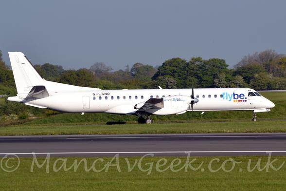 Side view of a white, twin propellor-engined airliner taxiing from left to right off the runway.
The plane is almost entirely white, with blue "FlyBe" titles above a series of yellow, red, and purple dots on the forward fuselage, just behind the cockpit.
There is the web address "www[dot]flybe[dot]com" in blue on the sides of the engine cowlings.
The registration "G-LGNR" is on the upper rear fuselage, in black.
The last 2 letters "NR" are on the front of the nose wheel doors, and above the cockpit corner windows, also in black.
Green grass lines either side of the black runway in the foreground, with the taxiway this plane is on hidden by grass.
More grass in the background leads up to trees in the distance, under blue-grey sky.