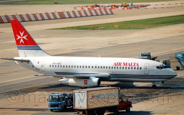 Side view of a white, twin engined jet airliner taxiing from left to right.
The plane has 3 thin blue stripes running along the body,  on a level with the wing root, and red "Air Malta" titles on the upper forward fuselage.
The registration "9H-ABG" is on the upper rear fuselage in black.
The bottom of the tail is white, with3 thin blue stripes at the top of that section, while the top is red with a white Maltese Cross (4 arrow-heads with the tips meeting in the middle.)
A smaller version of the same logo (white Maltese Cross on a red background over 3 blue lines) is on  the side of the engine pods.
Grey concrete apron fills most of the frame, with a white van and a blue fuel truck in the middle of the foreground.
More blue trucks and a blue set of mobile air stairs are in the background on the left.
A strip of green grass separates 2 taxiways, with a red and white striped metal fence and a small building site separating them from another in the distance.