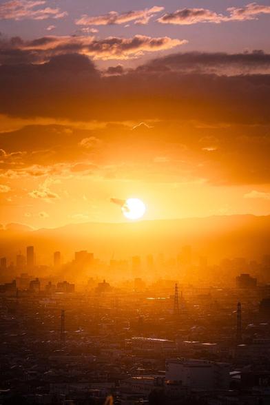 Amber light streams through buildings from a low sun above Osaka, as its bottom edge touches the horizon just before sunset.