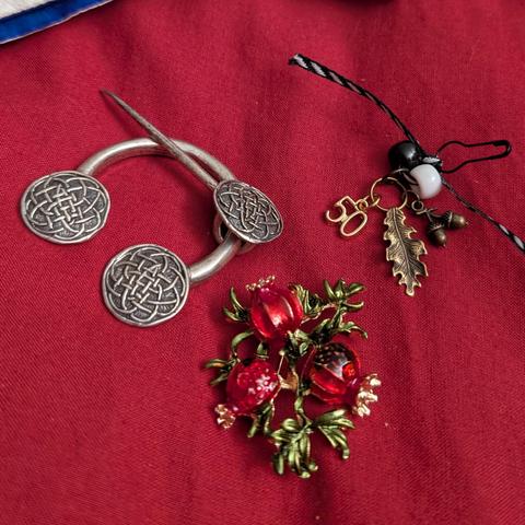 One cloak clasp with three circles of Celtic knotwork, a pin with three pomegranates, and a charm pin made from black/white diagonal striped ribbon, a black bead and a white bead, a charm that says 50, an oak leaf charm, and a collection of acorns charm