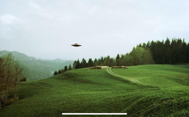 Photo of a UFO flying over a green landscape with wooded hills in the background. The UFO is distant in front of a bright but cloudy sky.